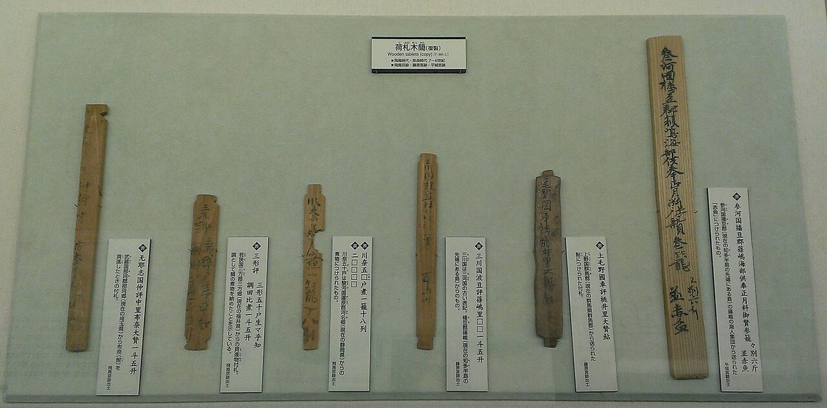 Mokkan, wooden tags inscribed in ink, shown as museum replicas. They were attached to shipments of tax goods and name the sender's province, district and village and the contents. The originals date from the 7th–8th centuries and were excavated at the palace sites of Asuka, Fujiwara and Nara (Heijō).