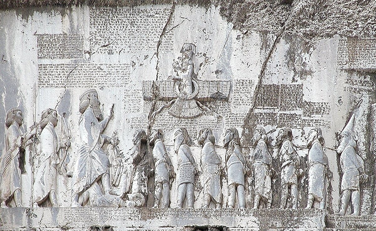The relief above the Behistun inscription (Kermanshah province, western Iran): Darius I, holding a bow, sets his foot on the defeated Gaumata; two attendants stand behind him, nine bound rebels before him, and above floats a winged figure usually interpreted as Ahura Mazda. Panels of cuneiform text surround the scene.